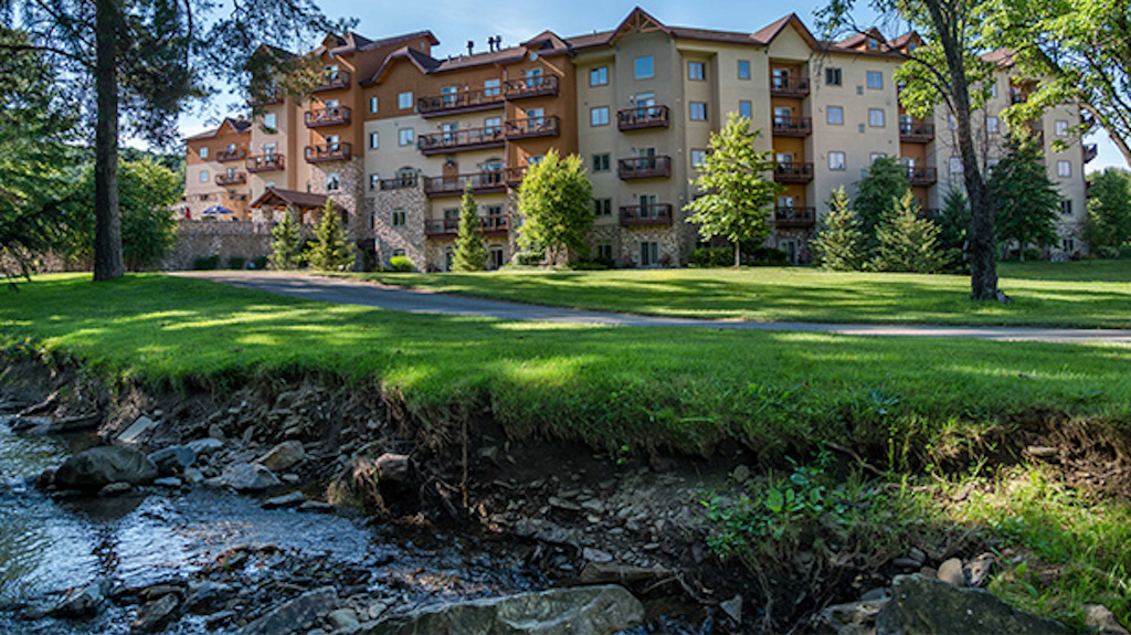A summertime exterior view of the Tamarack Club on a sunny day with a small creek in the foreground. 